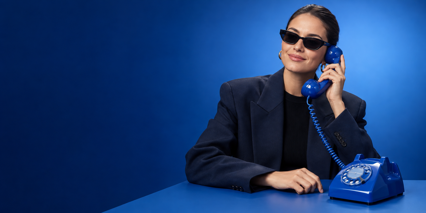 A woman in dark tailoring and sunglasses listening on a glossy blue telephone with a relaxed smile against a blue studio backdrop